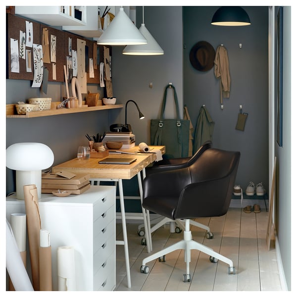 Home office with TOSSBERG black leather swivel chair. Wooden desk, shelves with art supplies, green walls, and hanging coats.