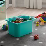 Turquoise TROFAST storage bin filled with toys on a gray carpet.