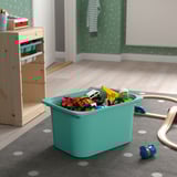 Turquoise toy bin filled with colorful toys on a grey polka dot rug, beside a wooden storage unit.