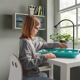 Child organizing tray at white desk with plant and lamp.