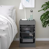 Gray TROFAST storage unit beside bed, holding lamp, water bottle, and baskets, with a white bed and plant nearby.