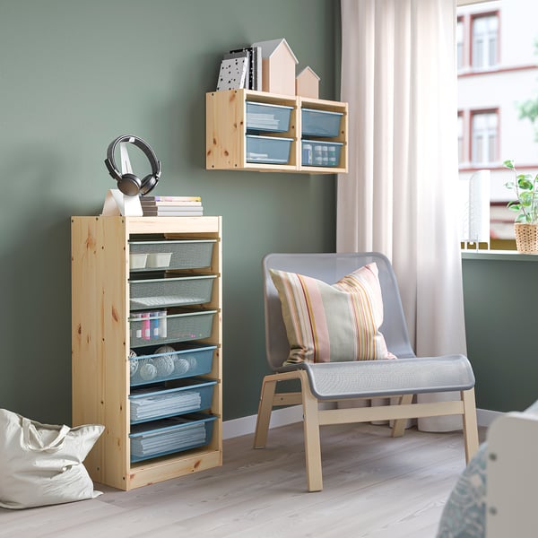 Wooden TROFAST unit with mesh drawers, holding books and headphones, paired with a cushioned chair and wall shelf for organization.