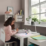 Girl sitting at white desk using white storage tray for arts and crafts. Desk and tray against purple wall, near window with plants.