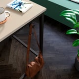 Brown leather bag hangs under desk; white mug, book, and green plant on desk.