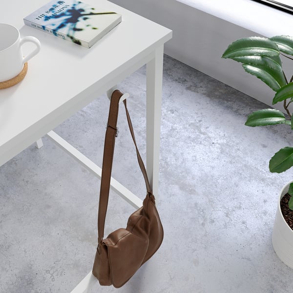 Brown leather bag hangs on white metal table leg. Table has white cup and book. Plant beside.
