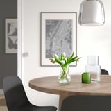 A dining table with a vase of white tulips, a green candle, and glass vases. Artwork and a gray pendant lamp hang above. Black chairs surround.
