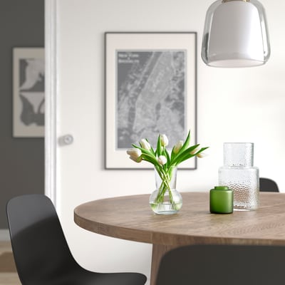 A dining table with a vase of white tulips, a green candle, and glass vases. Artwork and a gray pendant lamp hang above. Black chairs surround.