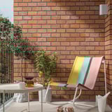 Modern colored rocking chair TUMHOLMEN against brick wall, near plants and table.