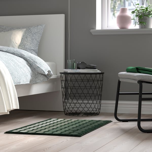 Cozy bedroom with dark green TUNNELBANA mat, patterned pillows, black side table, and plant.