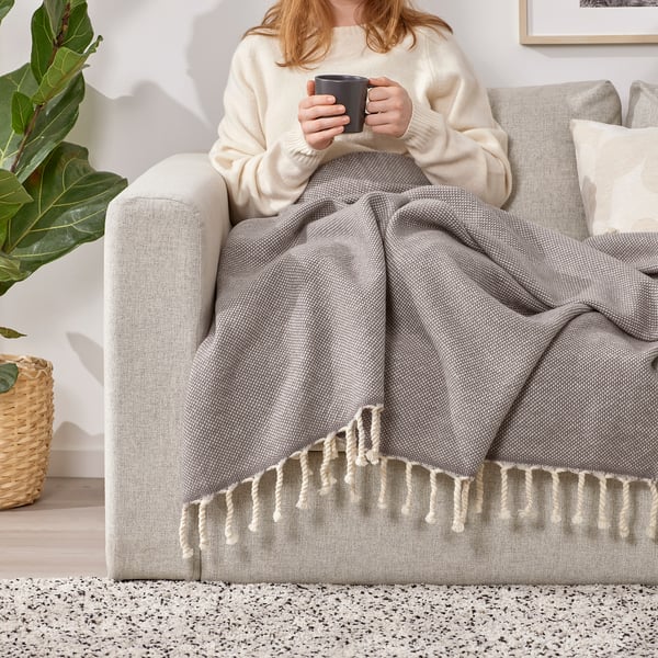 Person sitting on a grey couch, wrapped in a grey wool throw with fringe, holding a black mug.