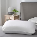 Large white pillow on bed, beside lamp, plant, small device, gray headboard.