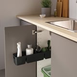 Black organizing bin mounted inside cabinet, holding cleaning spray, brushes, and bottle, mounted near kitchen sink.