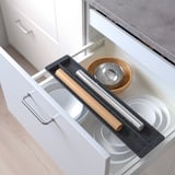 Open kitchen drawer with sliding organizer holding plates, rolling pin, and bowls. Organizer creates extra storage and dampens sound.
