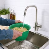 Person rinsing cooked pasta in green UPPFYLLD colander under stainless steel faucet.