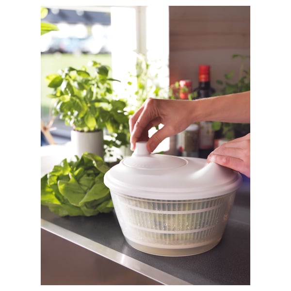 Person using UPPFYLLD salad spinner on kitchen counter, drying lettuce by spinning the lid. Fresh greens and plants in background.