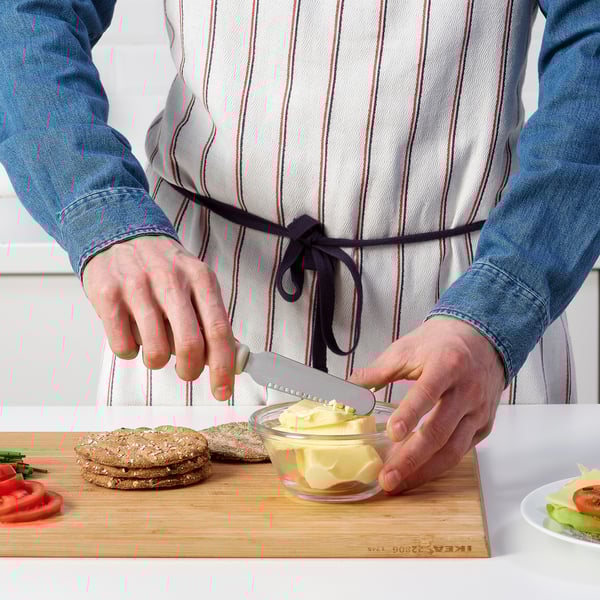 Person using UPPFYLLD butter knife on cutting board with bread and veggies.