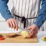 Person using UPPFYLLD butter knife on cutting board with bread and veggies.
