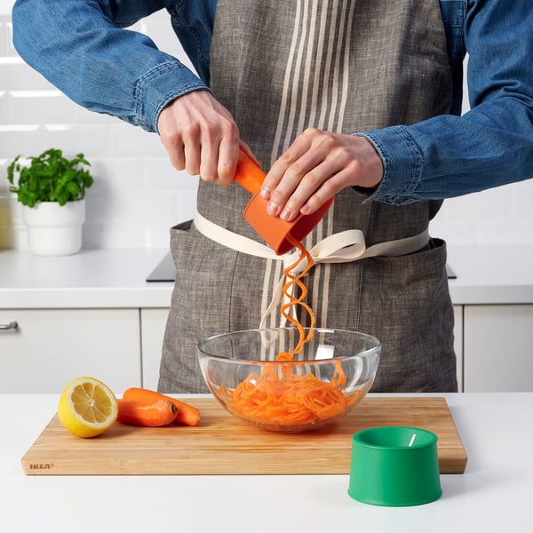 Person peels carrots using orange vegetable slicer, creating long strips in bowl. IKEA UPPFYLLD kitchen tool.