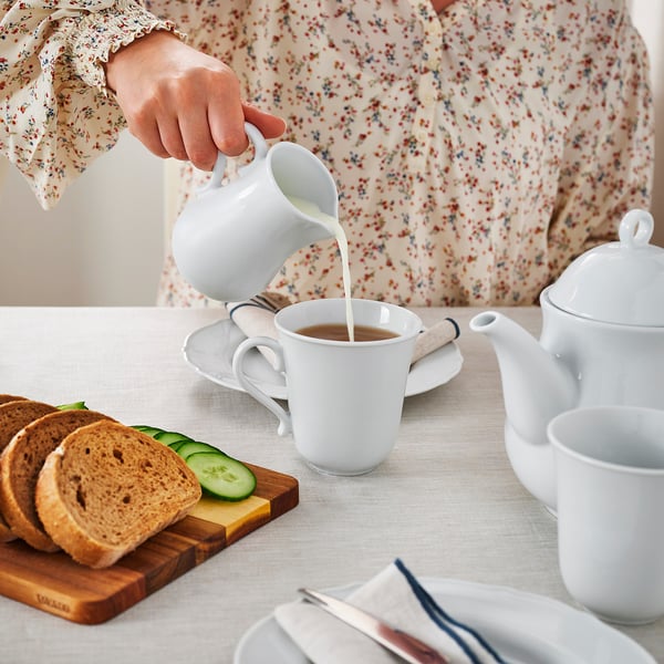 Woman pouring creamer into teacup. Table set with teapot, bread, cheese, cucumbers, and porcelain dishware.