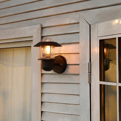 Black UPPLID outdoor wall lamp with clear glass shade, affixed to grey wooden wall, glowing warm light, small compared to surrounding structures.