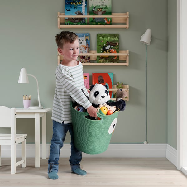 Boy holding green toy basket with stuffed panda, standing in a childs room with bookshelves and a desk.