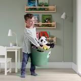 Boy holding green toy basket with stuffed panda, standing in a childs room with bookshelves and a desk.