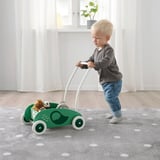 Blonde toddler standing on gray-speckled rug, pushing green dinosaur-themed walker with stuffed animals.
