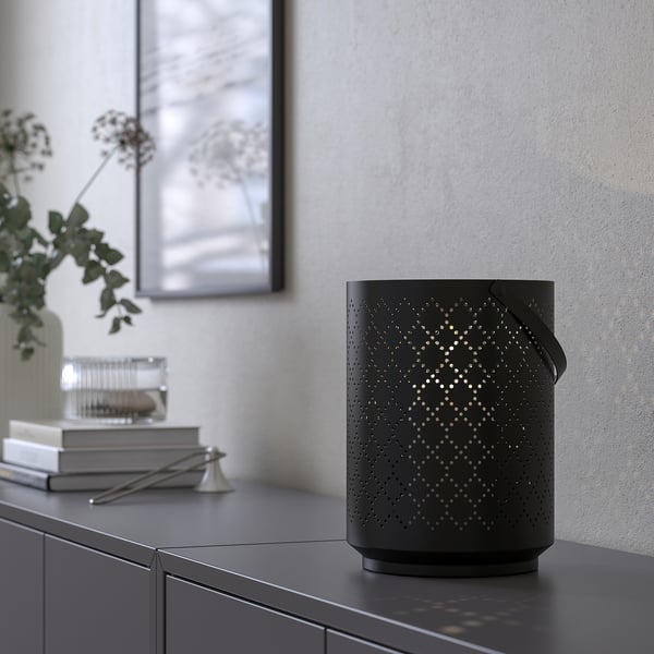 Black patterned lantern on shelf, casting light, with books, scissors, and vase nearby.