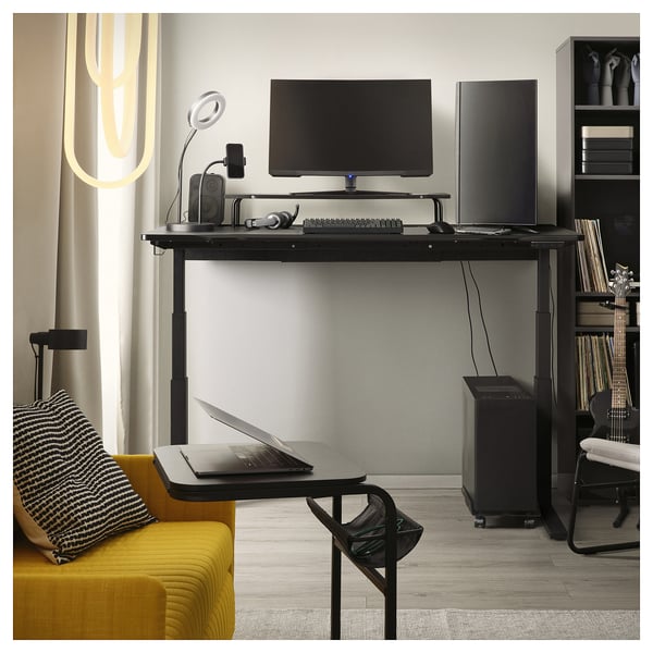 A black UTMANING adjustable gaming desk with a monitor, keyboard, laptop, and speakers. A yellow couch with a pillow is on the left side.