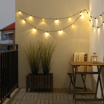 UTSUND outdoor lights curve above cozy balcony with plants & chairs, emitting warm glow.