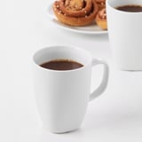 Two white ceramic mugs filled with coffee on a white table, accompanied by a plate of cinnamon buns.