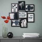 Modern gallery wall with black-framed abstract art prints. A vase with red flowers sits below on a shelf, alongside a stack of books.