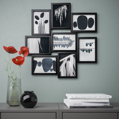 Modern gallery wall with black-framed abstract art prints. A vase with red flowers sits below on a shelf, alongside a stack of books.