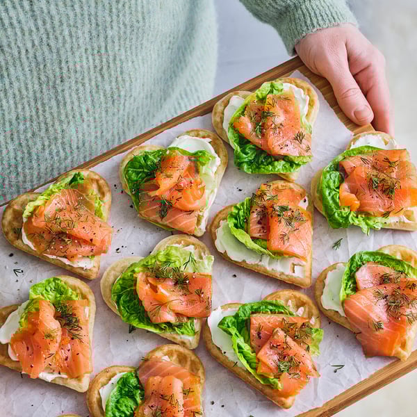 Heart-shaped waffle appetizers topped with smoked salmon, lettuce, and dill. Ready-baked and served on a wooden board.