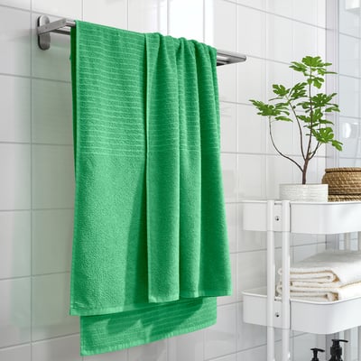 Green VÅGSJÖN towel on chrome bar, above white towels, vibrant against tiles, plant adds nature.