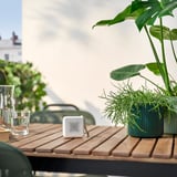 Small white VAPPEBY speaker sits on wooden table, surrounded by plants and drink glasses. Compact and portable.