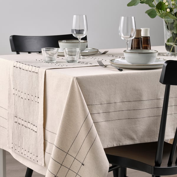 Dining table with beige runner, subtle patterns, tassels, black chairs, glassware, ceramic dishes for cozy meal.