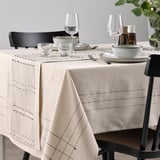 Dining table with beige runner, subtle patterns, tassels, black chairs, glassware, ceramic dishes for cozy meal.