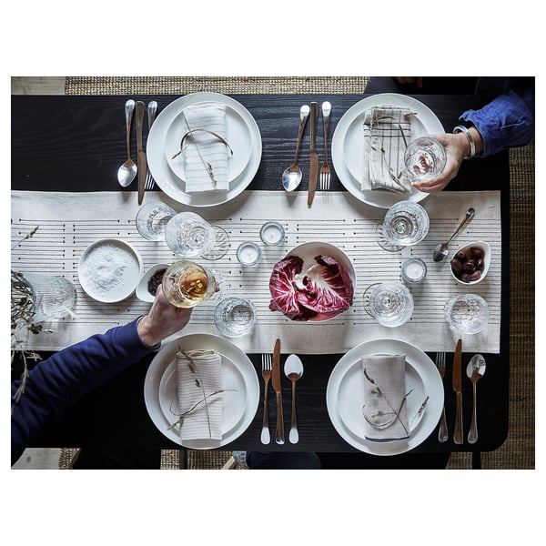 Four people at a dark wooden table with the VÅRARV table runner, white plates, wooden utensils, glasses, and a large leafy vegetable.