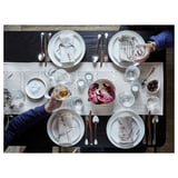 Four people at a dark wooden table with the VÅRARV table runner, white plates, wooden utensils, glasses, and a large leafy vegetable.