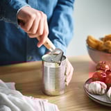 Person opens can using VARDAGEN bottle/can opener on wooden table, with tomatoes and garlic nearby.