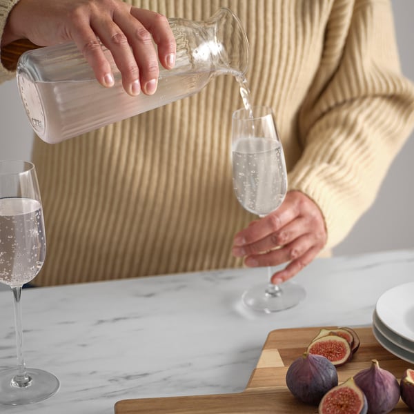 Person pouring water from clear glass carafe into a wine glass, showcasing VARDAGEN glasswares versatility for serving beverages.