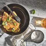 Dark frying pan with food, knife, fork, salt bowl, and drink on marble table.