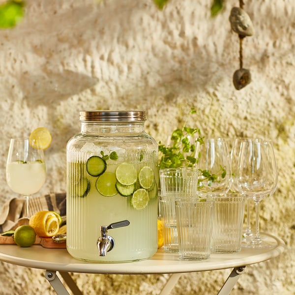 Clear glass drinks dispenser with silver tap, lime slices, glasses, on wooden stand against stone wall.