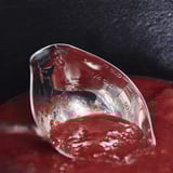 Clear measuring ladle tipped with red liquid, markings visible, on black and red surface.