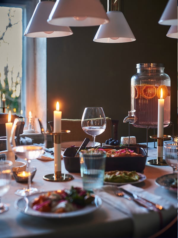 Lit dining table with plates, glasses, candles, and a VARDAGEN jar stand in silver, showcasing elegant dinner setup.