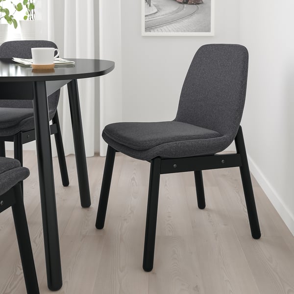 Modern black VEDBO chair with dark grey upholstery, curved back for support, sits beside a small table with a cup, in a well-lit room.
