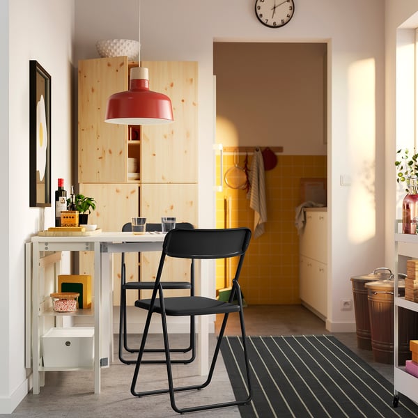 Modern kitchen with black folding chairs, wooden table, red pendant lamp, and shelves. Yellow-tiled kitchen with white walls.