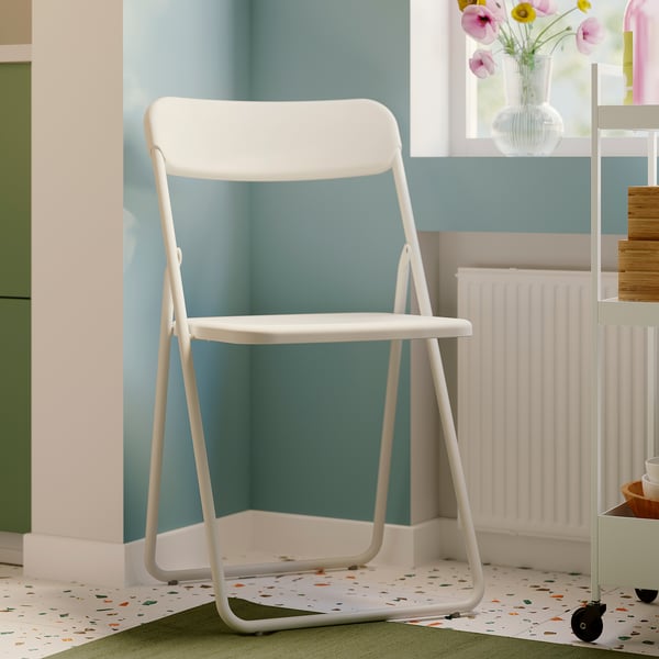 White VIHALS folding chair, slim & modern, sits neatly in corner by window, fits well with nearby radiator & shelves.