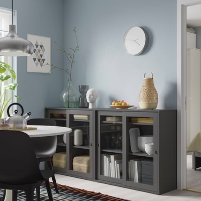 Grey storage unit with glass doors, clock above, holds dishes & books, next to black chair.